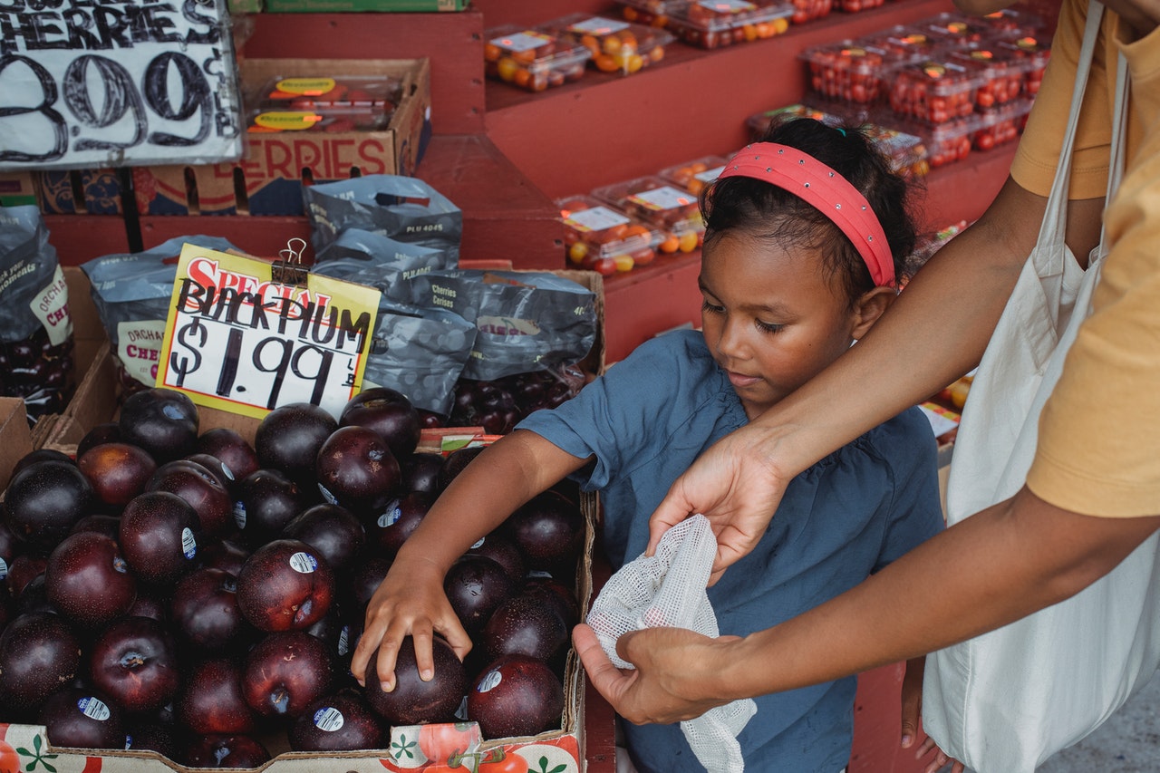 Little girl grabbing plums at grocery stand with mother.